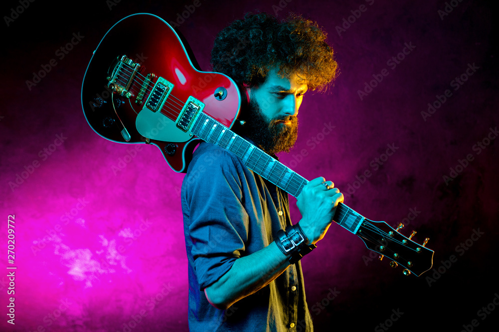 Obraz premium Portrait of hipster man with curly hair with red guitar in neon lights. Rock musician is playing electrical guitar.