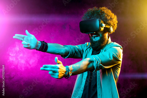 Photo of handsome bearded hipster man with curly hair shooting virtual guns with goggles in neon lights. Smartphone using with VR headset.