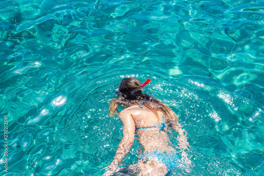 swimming girl in snorkeling mask and tube looking underwater on bottom through transparent vivid blue Red sea water back to camera, foreshortening from above, empty copy space for text