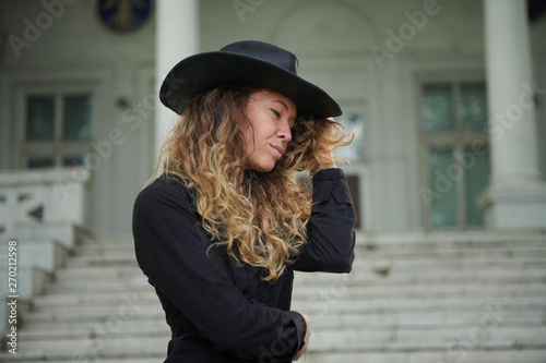 fashionable girl dressed in black shirt, hat and wide trousers posing near old white house