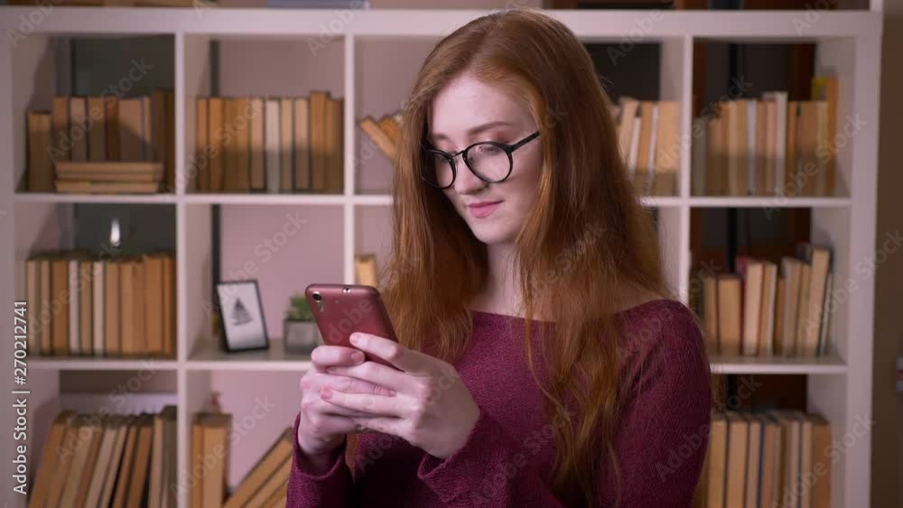 Closeup portrait of young redhead attractive caucasian female student in glasses using the phone smiling happily in the college library Closeup portrait of young redhead attractive caucasian female student in glasses using the phone smiling happily in the college library