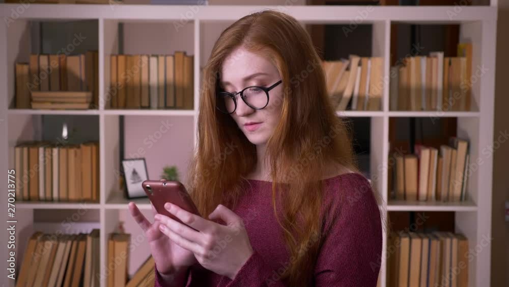 Closeup portrait of young redhead attractive caucasian female student in glasses texting on the phone in the college library Closeup portrait of young redhead attractive caucasian female student in glasses texting on the phone in the college library