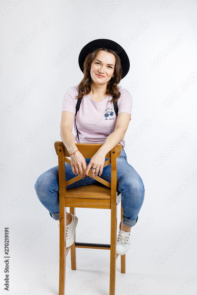 Young beautiful girl sitting on wooden chair in stylish black hat on white background. Emotions and gestures. Young beautiful girl sitting on wooden chair in stylish black hat on white background. Emotions and gestures.