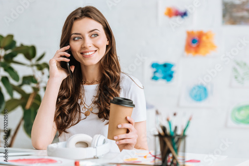 cheerful girl talking on smartphone and holding paper cup while sitting at table near headphones
