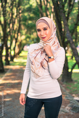 Young attractive Muslim Malay woman, modern contemporary fashion in hijab, close up portrait face and body, blurred background smiling and happy at park.