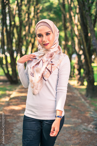 Young attractive Muslim Malay woman, modern contemporary fashion in hijab, close up portrait face and body, blurred background smiling and happy at park.