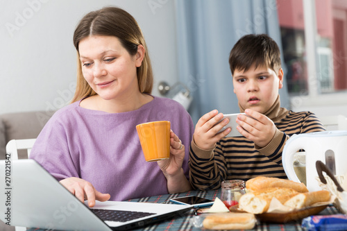 Young woman working at laptop and son using phone at table with tea