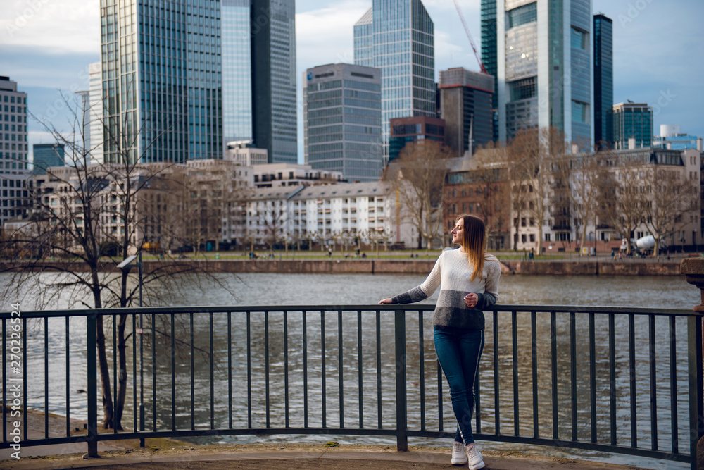 Beautiful young girl with long straight hair standing near skyscrapers and river in Frankfurt am Main, Germany
