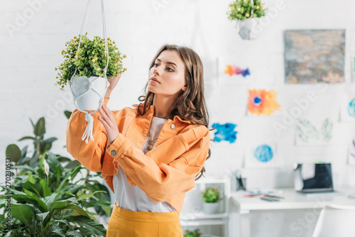 beautiful woman in orange clothing touching hanging flowerpot at home