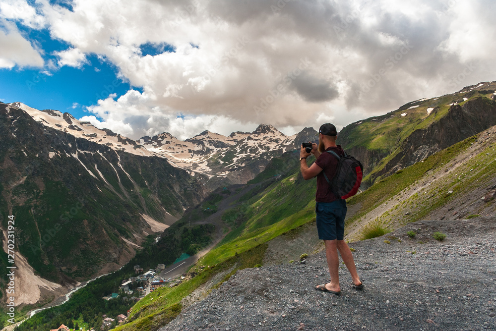 Tourist with backpack stands on top of plateau and photographs mountains. Caucasus Mountains, Gorge Baksan