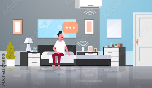 man sitting on bed using conditioner and clock controlled by smart house speaker voice recognition concept modern bedroom interior flat horizontal full length