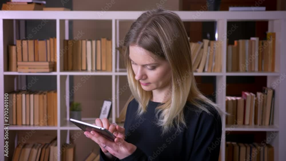 Closeup shoot of young attractive female student using the tablet looking at camera smiling happily indoors in the university library Closeup shoot of young attractive female student using the tablet looking at camera smiling happily indoors in the university library