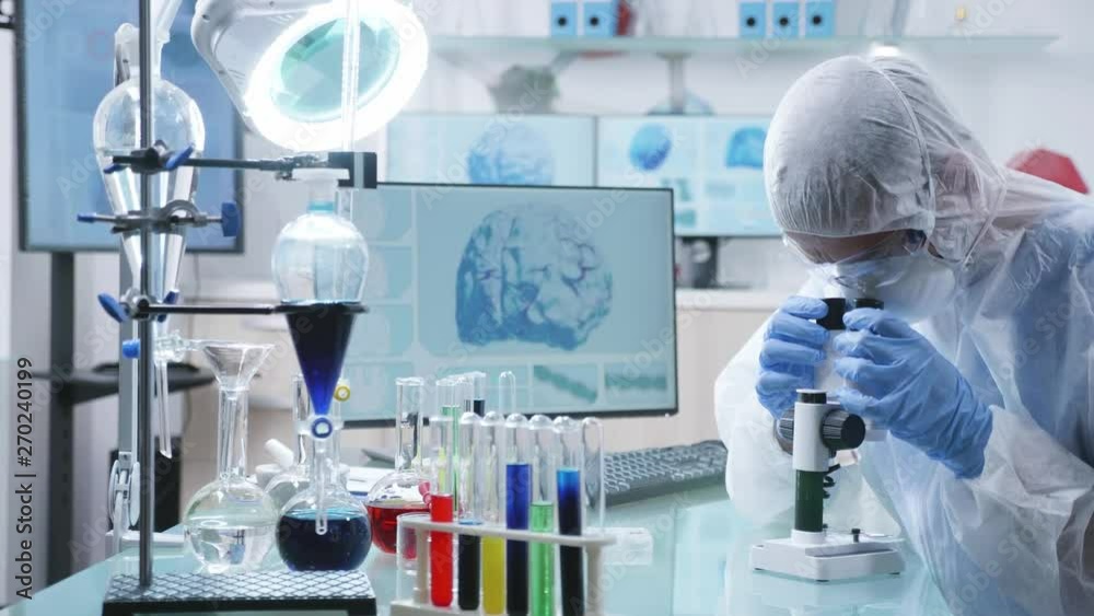Zoom in shot of scientist researcher in professional high end laboratory looking into the microscope. Various colored liquids are in tubes on the table Zoom in shot of scientist researcher in professional high end laboratory looking into the microscope. Various colored liquids are in tubes on the table