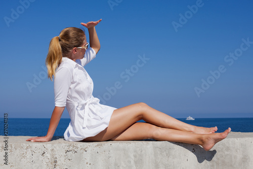 Barefoot girl in white short dress sits leaning on hand and looking away with hand visor