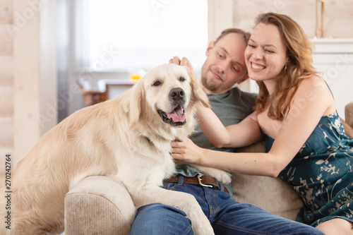 Young happy married couple nice girl and young man in casual clothes talking with his kind obedient dog in the living room of his cozy country house © Rithor