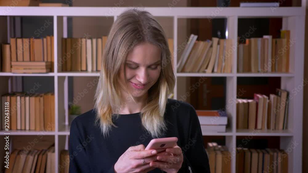 Closeup shoot of young attractive female student using the phone looking at camera smiling happily indoors in the university library Closeup shoot of young attractive female student using the phone looking at camera smiling happily indoors in the university library