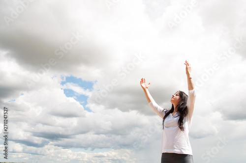 a woman stands with her arms raised and prays to God. On a green field in summer. Blue sky and white clouds.