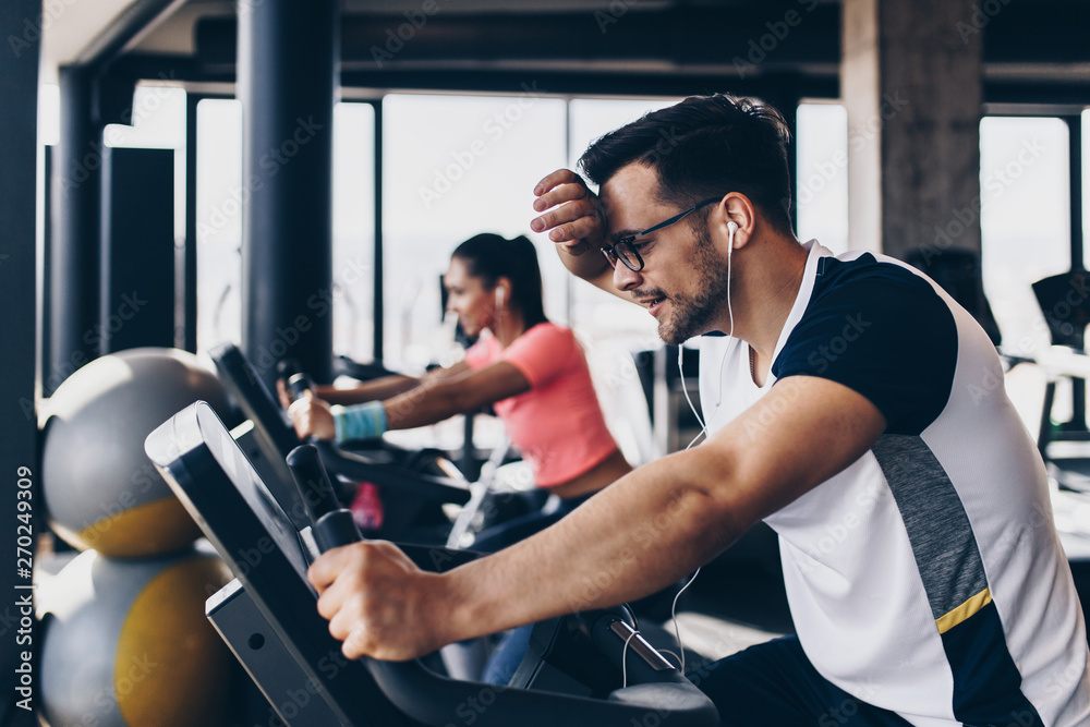 Young and attractive woman and man biking in the fitness gym. They exercising legs and doing cardio workout while riding cycling machines.