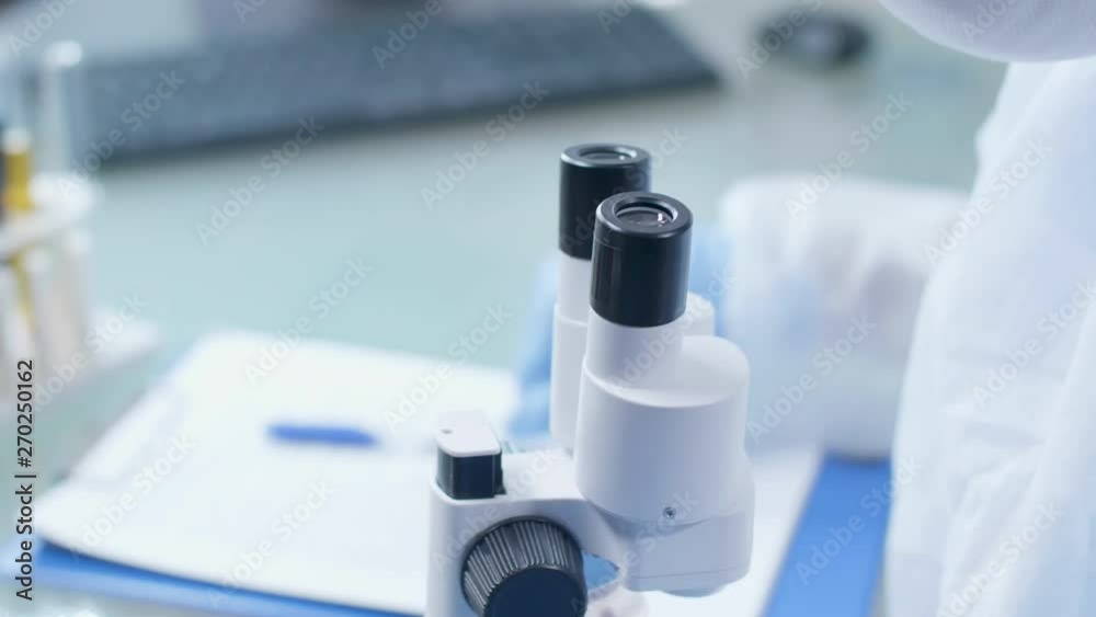 Close up handheld shot of lab technician taking notes while looking into the microscope. Science and chemistry Close up handheld shot of lab technician taking notes while looking into the microscope. Science and chemistry