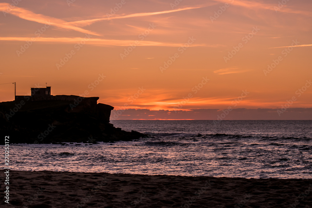 Silhouette of fort and cliff with colorful and stunning sunset in the background, dusk at São Lourenço beach, Ericeira - Mafra, Portugal