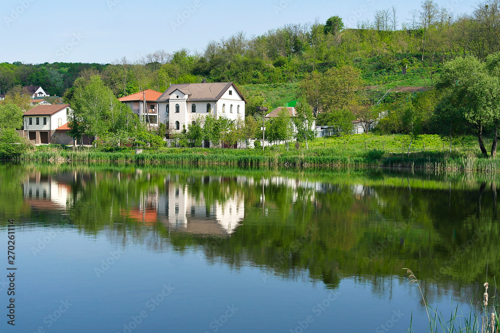House under the hill on the lake. Clear summer day. Luxurious reflection in the lake.