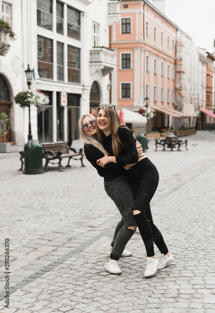 Happy friends having fun walking in city center. Beautiful young positive girls spend summer holidays. Two smiling pretty teenagers travel around old wonderful European city. Joint vacation concept