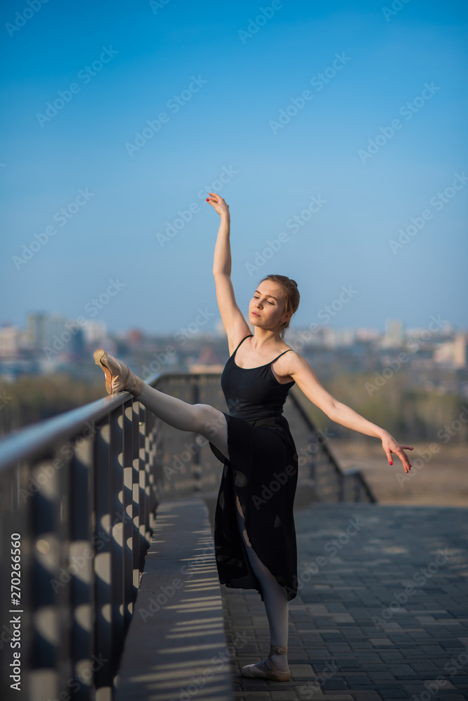 Ballerina in a tutu posing near the fence. Beautiful young woman in black dress and pointe dancing outside. Gorgeous ballerina demonstrates amazing stretching.