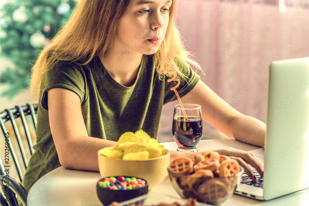 Girl works at a computer and eats fast food. Unhealthy food: chips, crackers, candy, waffles, cola. Junk food, concept. Girl works at a computer and eats fast food. Unhealthy food: chips, crackers, candy, waffles, cola. Junk food, concept.