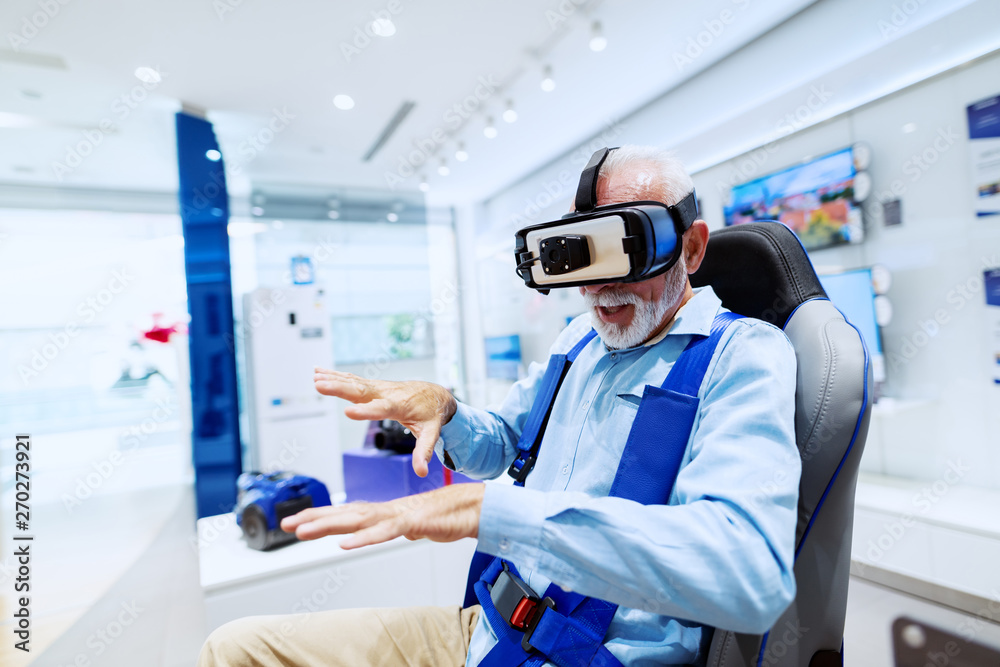Excited bearded senior man sitting in chair and trying out virtual reality goggles. Tech store interior. Excited bearded senior man sitting in chair and trying out virtual reality goggles. Tech store interior.