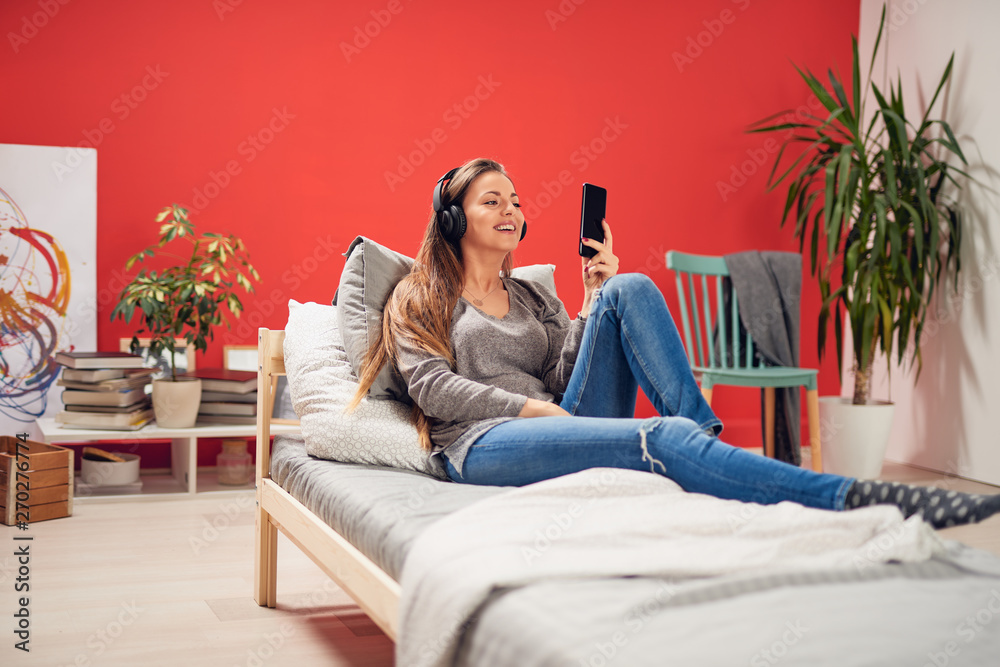 Obraz premium Young Caucasian brunette sitting on the bed with headphones on ears and using smart phone. Coral red background.