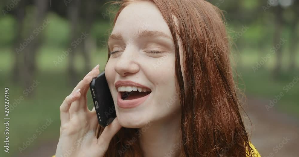 Young, attractive redhead girl talking on the phone while standing in the rain. The girl is very happy to communicate. in slow motion. Shot on Canon 1DX mark2 4K camera Young, attractive redhead girl talking on the phone while standing in the rain. The girl is very happy to communicate. in slow motion. Shot on Canon 1DX mark2 4K camera