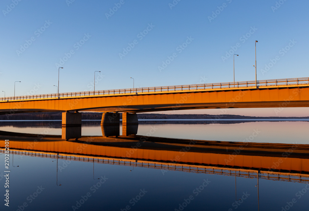 Long bridge over calm lake during golden hour. Orange glowing concrete bridge during sunset.