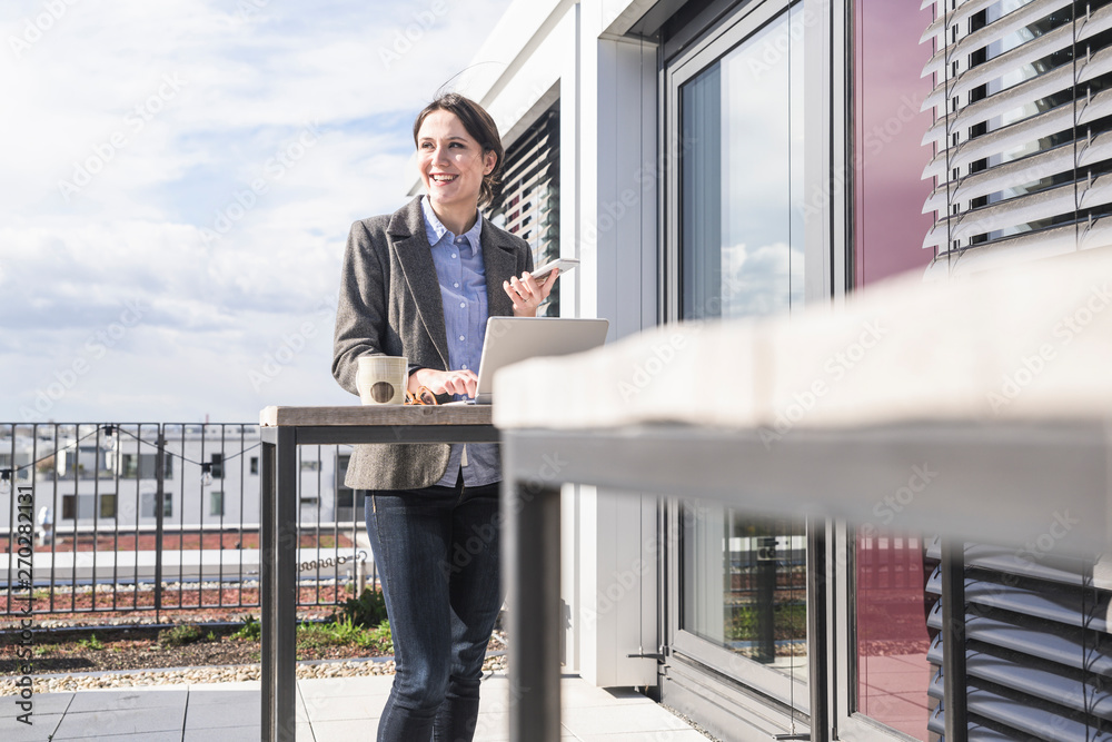 Smiling businesswoman with cell phone and laptop on roof terrace Smiling businesswoman with cell phone and laptop on roof terrace