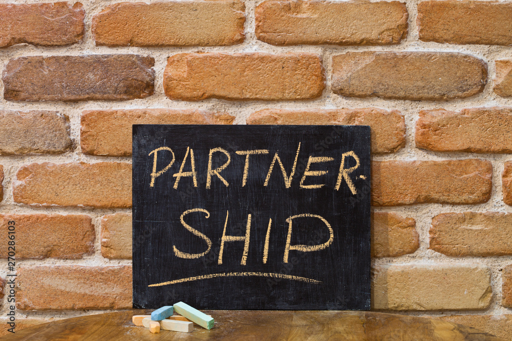 Fototapeta premium Chalk board with the word PARTNERSHIP drown by hand and chalks on wooden table on brick wall background.