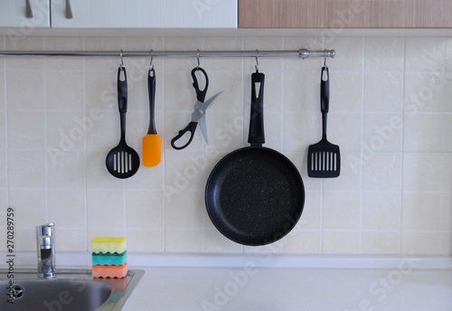 Non-stick frying pan, scissors for food, shovels for turning steaks, a spoon against the background of a tiled wall  .The interior of the kitchen.
