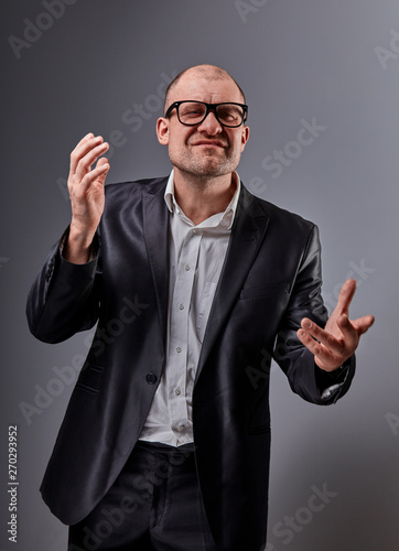 Fototapet Thinking angry bald business man in eyeglasses and suit discussing and moving the hands  on grey background