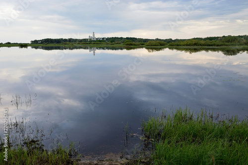 A lake with green grass in the foreground and a reflection of the cloudy sky and on the horizon a Christian monastery.Yuryev Monastery Veliky Novgorod.Lake Myachino Novgorod region