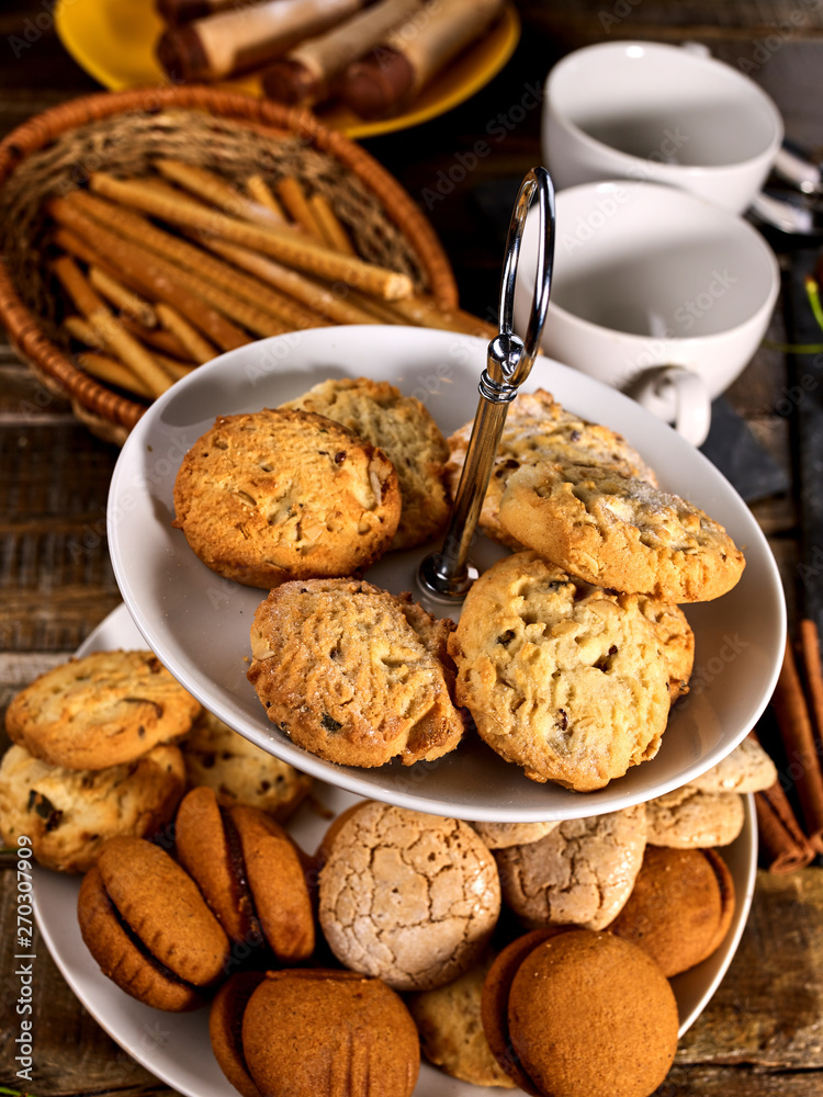 Oatmeal and nut cookies and sand biscuit on tier cake stand with straw pastry in basket on kitchen on wooden table in rustic natural style . Top view of two cups of tea on wooden table.
