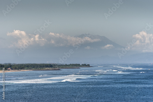 Bali, Indonesia - February 25, 2019: Ji Pantai Serangan beach north of Benoa Harbour Entrance. Blue water, sandy beach, green forest belt, tall mountain partly hidden in white clouds over light blue s