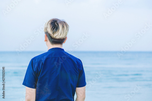 The young man wears a blue shirt. Standing on the sand by looking at the sea