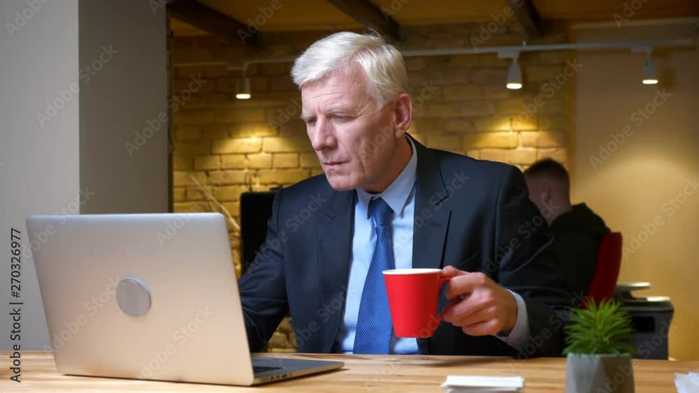 Closeup shoot of old caucasian businessman using the laptop and drinking coffee being busy and focused indoors in the office on the workplace Closeup shoot of old caucasian businessman using the laptop and drinking coffee being busy and focused indoors in the office on the workplace