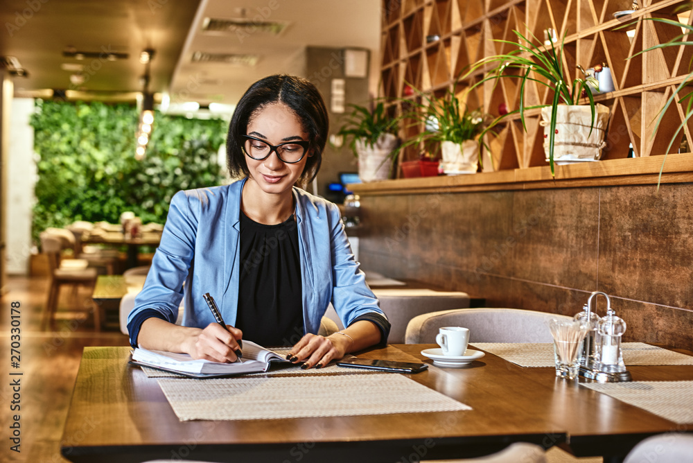The greatest ability in business is to get along with others and to influence their actions. Young beautiful businesswoman smiling, writing in notebook while working in cafe The greatest ability in business is to get along with others and to influence their actions. Young beautiful businesswoman smiling, writing in notebook while working in cafe