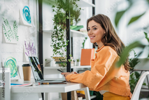 selective focus of smiling young woman using smartphone while sitting at desk near laptop