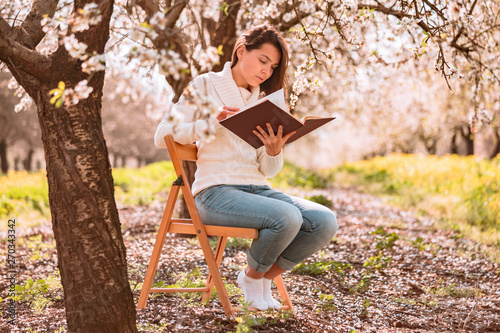 Young beautiful lady in white sweater sitting on wooden chair under blooming three and reading the book