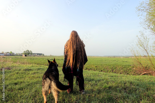 A girl with long flowing hair in a poncho and a German shepherd in the field