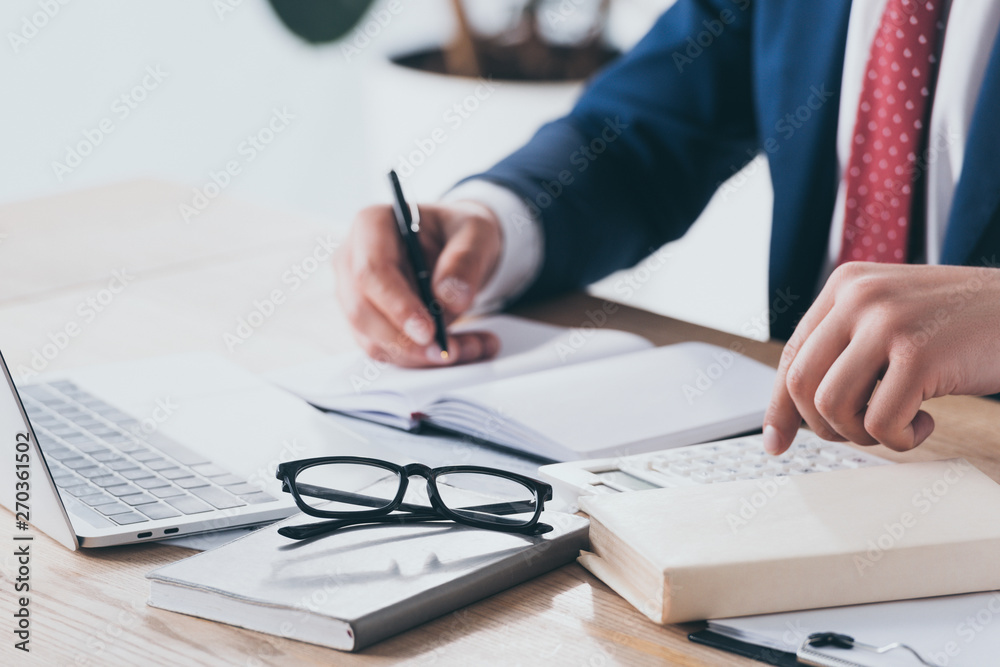 partial view of businessman in formal wear using calculator and writing in notebook at workplace partial view of businessman in formal wear using calculator and writing in notebook at workplace