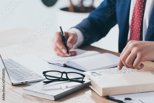 partial view of businessman in formal wear using calculator and writing in notebook at workplace