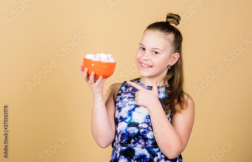 Kid girl with long hair likes sweets and treats. Calorie and diet. Hungry kid. Marshmallow challenge. Girl smiling face hold bowl with sweet marshmallows in hand beige background. Sweet tooth concept