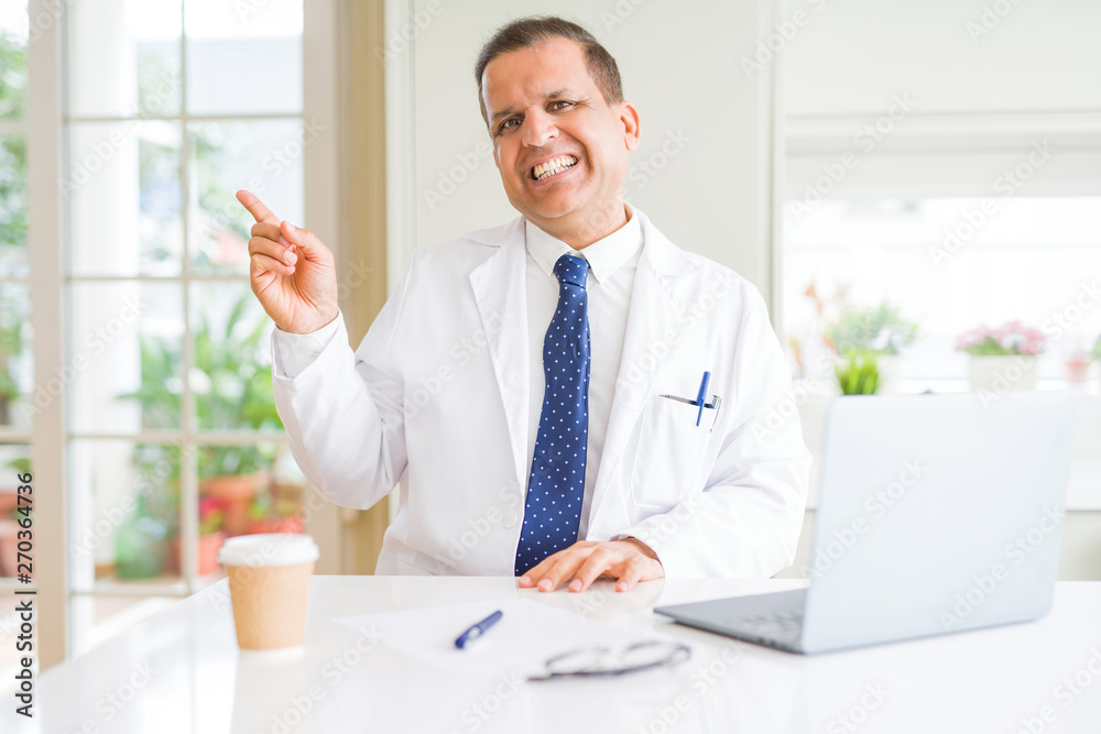 Middle age doctor man wearing white medical coat working with laptop at the clinic with a big smile on face, pointing with hand and finger to the side looking at the camera.