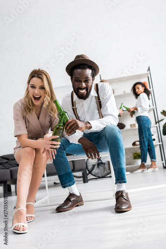 low angle view of happy girl laughing with cheerful african american man holding bottle of beer